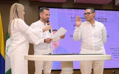 Posesión del rector en la Universidad Popular del Cesar con un hombre juramentando con la mano levantada frente a el presidente del consejo superior y una asistente, todos vestidos de blanco.